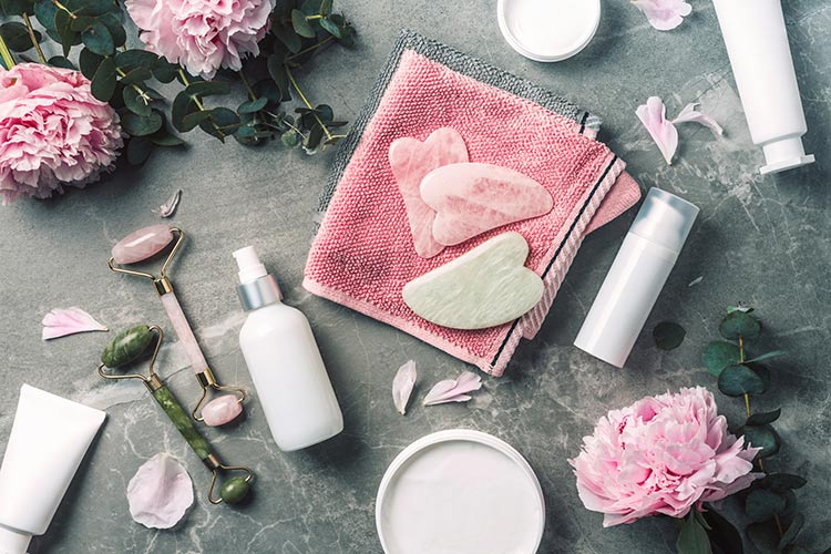 cosmetic-products-bottles-towels-oil-face-roller-gua-sha-massager-pink-peonies-flowers